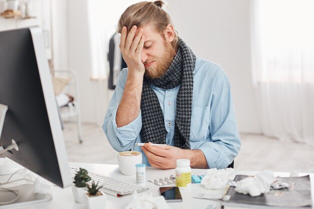 Sick bearded male in despair uses thermometer to measure temperature of body, sadly looks at it, suffers from high temperature, feels bad, surrounded by medicine and handkerchiefs at his workplace.