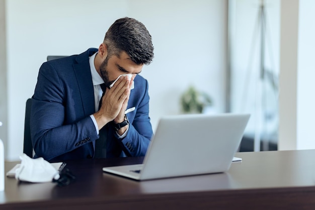 Free photo sick businessman sneezing in a tissue at his office desk