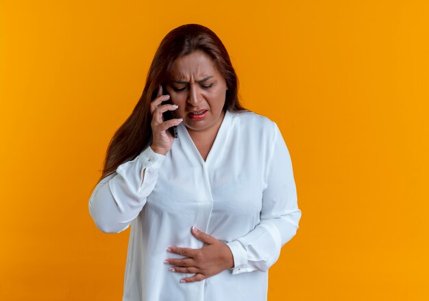 Sick casual caucasian middle-aged woman speaks on phone and putting hand on aching stomach isolated on yellow wall