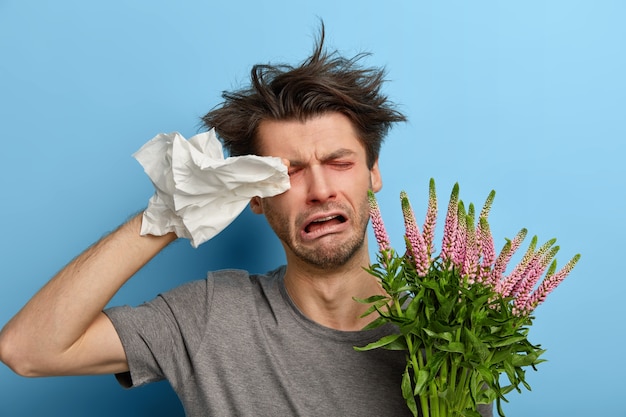 Sick dejected young man rubs eyes with handkerchief, has allergy to seasonal flowers or plants, cries unhappily, being tired of fighting against allergens, needs good treatments, stands indoor