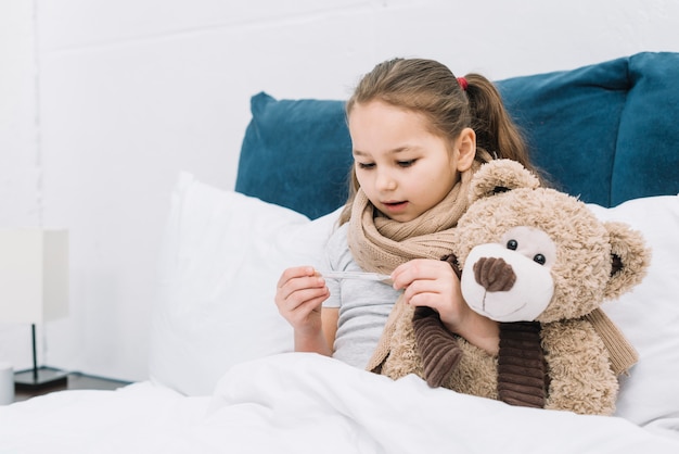 Sick girl sitting on bed with teddy bear looking at thermometer