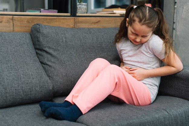 Sick girl sitting on gray sofa suffers from stomach pain at home
