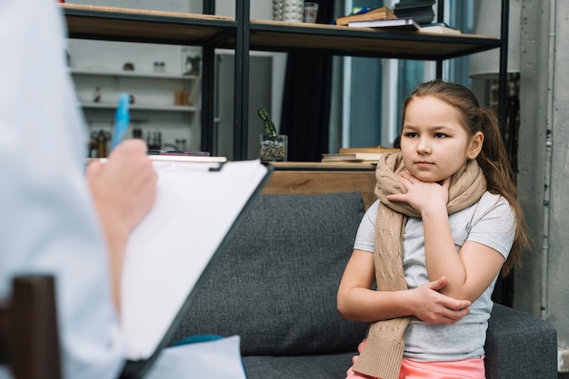 Free photo sick girl with scarf around her neck sitting in front of woman writing on clipboard with pen