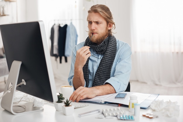 Больной больной бородатый мужчина сидит перед компьютером, пытается сосредоточиться на работе, держит в руке очки. Исчерпаны офисный работник устал, сидячий образ жизни, изолированных на фоне офиса.