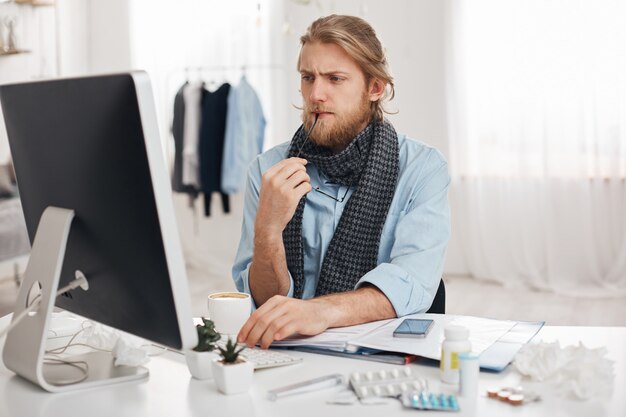 Sick ill bearded man sits in front of computer, tries to concentrate on work, holds spectacles in hand. Exahausted office worker tired, has sedentary lifestyle, isolated against office background.