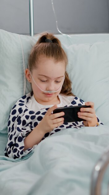 スマートフォンを使用してオンラインビデオゲームをプレイしてベッドで休んでいる病気の小さな子供