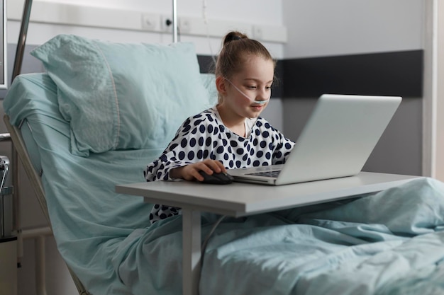 Sick little girl playing games on laptop while hospitalized in healthcare clinic pediatric ward room. Ill kid resting on patient bed while enjoying gaming on modern computer.