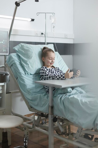 소아과 진료실에서 환자 침대에서 쉬면서 스마트폰으로 노는 아픈 어린 소녀. 병원 회복 병동에 앉아 있는 동안 휴대폰 엔터테인먼트를 즐기는 즐거운 젊은 환자.