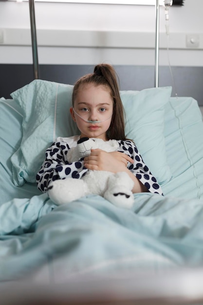 Sick little girl under treatment wearing oxygen tube while holding plush bear toy. Ill kid resting in children healthcare facility patient bed alone while having teddy bear.