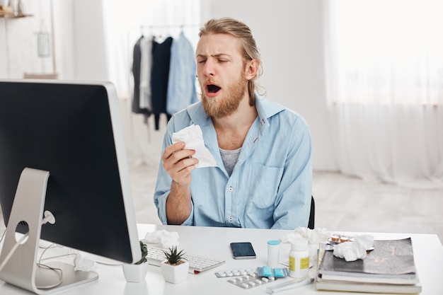 Sick male office worker holds handkerchief, sneezes, has unhappy and tired expression, isolated against office background. Unhealthy young man spreads bacteria