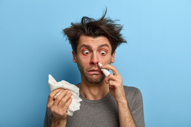 Free photo sick man injects nasal drop for blocked nose, suffers from cold, allergy or rhinits, rubs with handkerchief, feels unwell, has bugged eyes, isolated on blue wall stays at home during course of disease