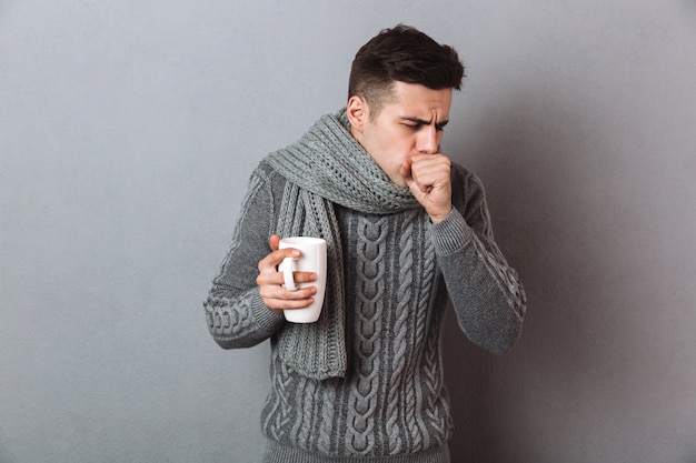 Sick Man in sweater and scarf holding cup of tea while having cough