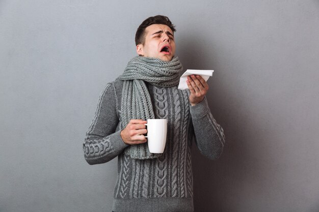 セーターとスカーフで病気の男がお茶を押しながらくしゃみをする