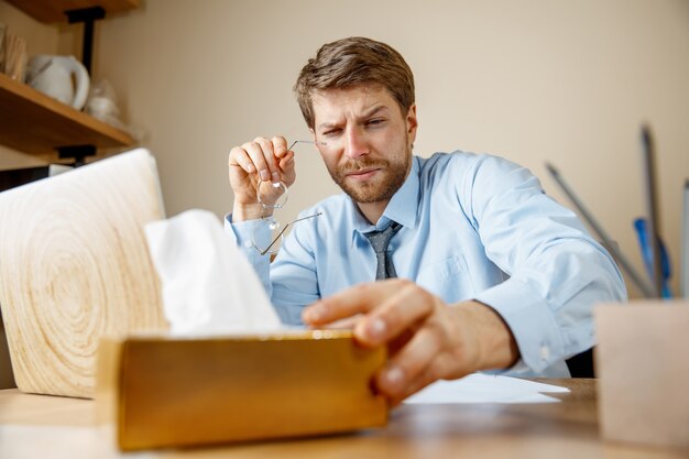 Sick man with handkerchief sneezing blowing nose while working in office, businessman caught cold, seasonal flu.
