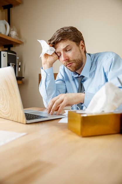 Sick man with handkerchief sneezing blowing nose while working in office