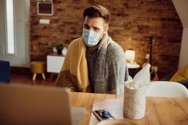 Больной мужчина, завернутый в одеяло, с помощью ноутбука дома во время пандемии коронавируса