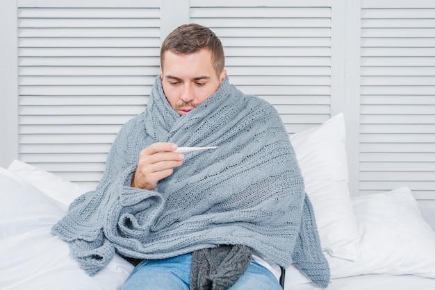 Sick man wrapped in shawl looking at thermometer