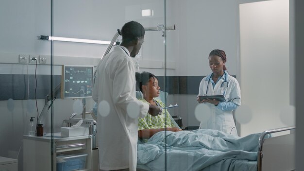 Sick patient receiving medical assistance from team of doctors in hospital ward. Specialists using digital tablet and checkup files to cure woman with disease in bed. Healthcare recovery