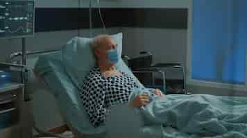 무료 사진 병원 병동 침대에 안면 마스크와 산소 농도계가 있는 아픈 환자가 앉아 있습니다. 건강 문제와 질병을 치료하기 위해 iv 드립 백에서 치료를 기다리는 질병을 가진 노인