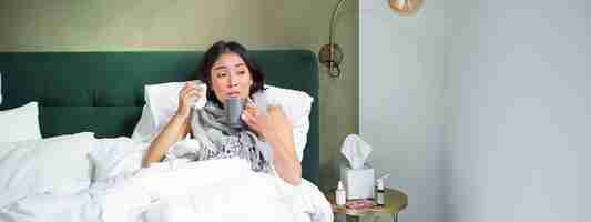 無料写真 インフルエンザでベッドに横たわる病気の悲しいアジアの女の子が風邪を引き、薬を飲み、熱いお茶を飲んでいる感じ