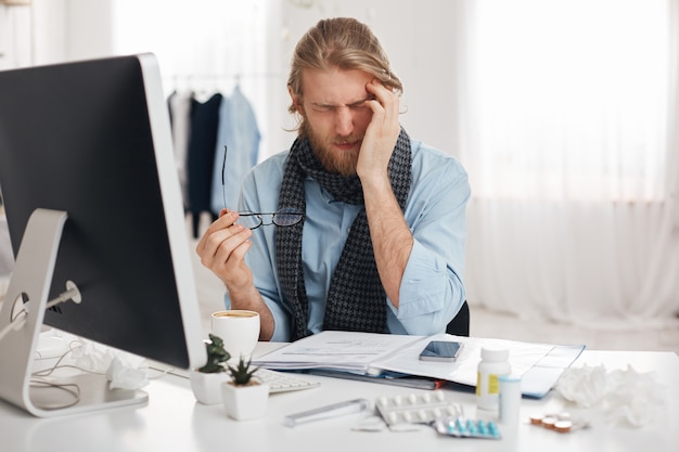 Free photo sick and tired bearded male student or office worker has sleepy expression, runs temple because of feeling sick, surrounded by pills and drugs, tries to concentrate and finish work quicker