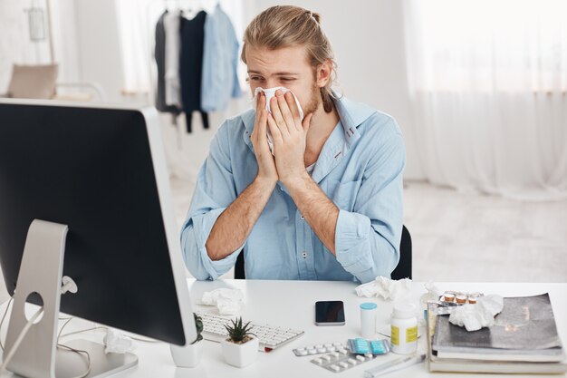 Sick and tired bearded office worker has suffering expression, has running nose, sneezing, coughing, because of flu, surrounded by pills and drugs, tries to concentrate and finish work quicker