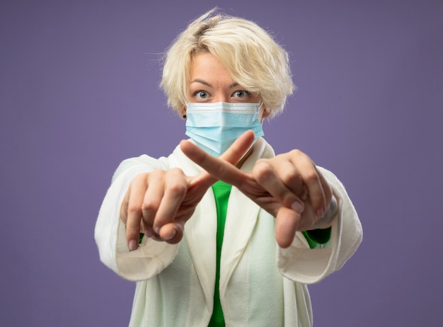 紫色の壁の上に立っている人差し指を横切る一時停止の標識を作る顔の保護マスクの短い髪の病気の不健康な女性