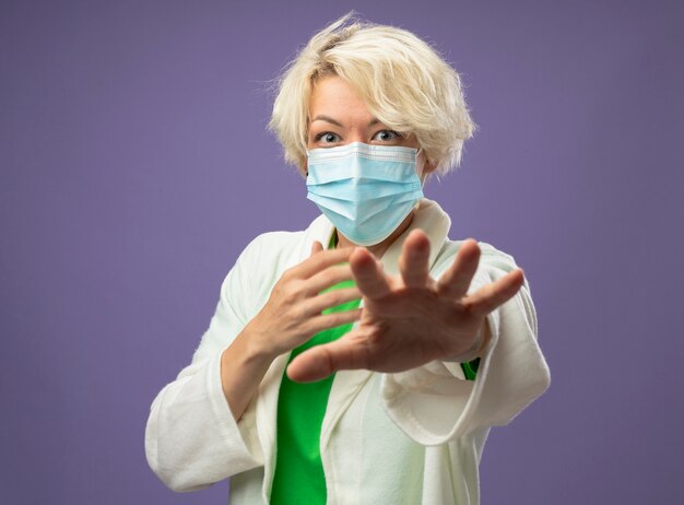 Sick unhealthy woman with short hair in facial protective mask  making stop sign with handstanding over purple wall