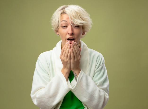 Sick unhealthy woman with short hair feeling unwell sneezing standing over light wall
