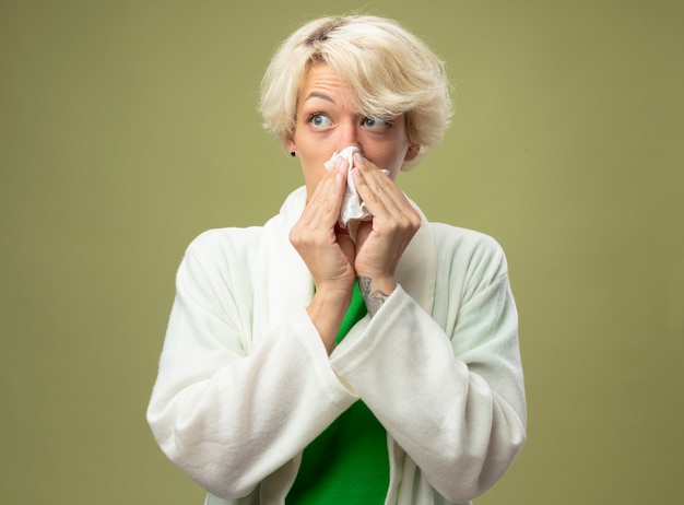 Sick unhealthy woman with short hair feeling unwell wiping her nose with tissue standing over light wall