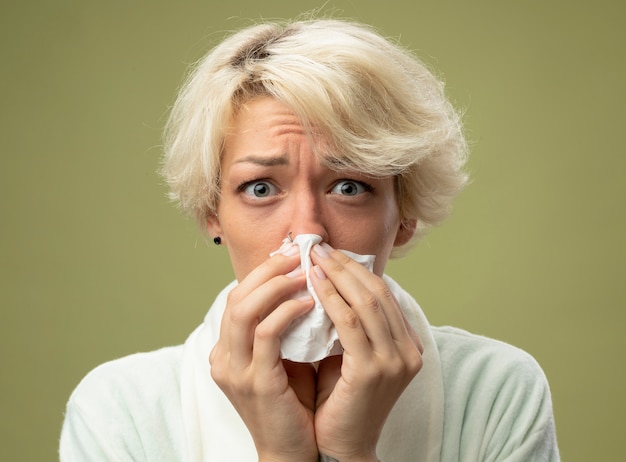 Sick unhealthy woman with short hair feeling unwell wiping nose with tissue standing over light wall