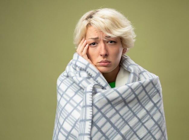 Sick unhealthy woman with short hair feeling unwell wrapped in blanket looking suffering from flu touching her head standing over light wall