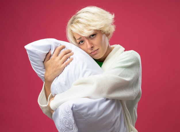 Sick unhealthy woman with short hair hugging pillow feeling unwell looking at camera with sad expression standing over pink background