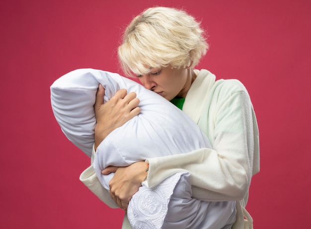 Sick unhealthy woman with short hair hugging pillow feeling unwell trying to smell it standing over pink background