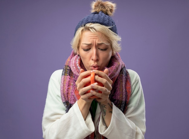 Бесплатное фото Больная нездоровая женщина с короткими волосами в теплом шарфе и шляпе плохо себя чувствует, держа чашку горячего чая, собираясь выпить, стоя на фиолетовом фоне