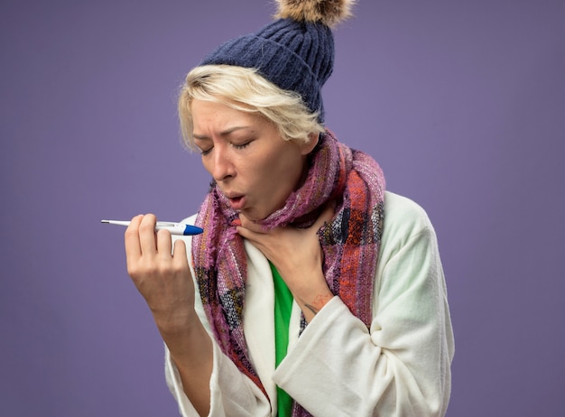 Бесплатное фото Больная нездоровая женщина с короткими волосами в теплом шарфе и шляпе плохо себя чувствует с термометром и кашляет, страдая от гриппа, стоя на фиолетовом фоне