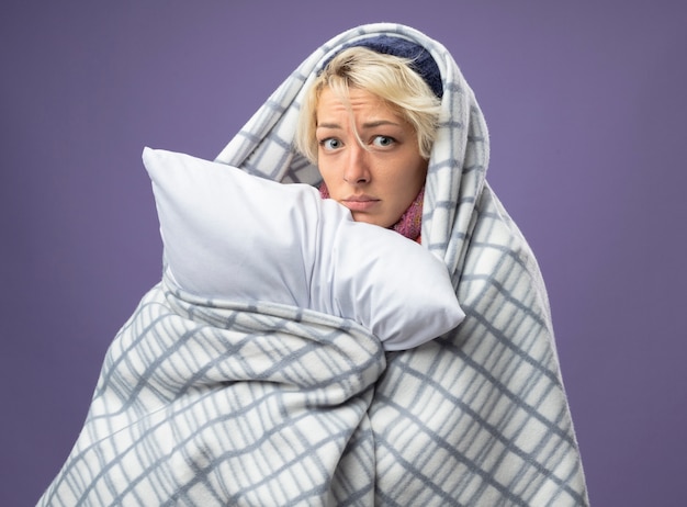 Free photo sick unhealthy woman with short hair in warm hat wrapped in blanket holding pillow feeling unwell unhappy and worried looking at cameraover purple background