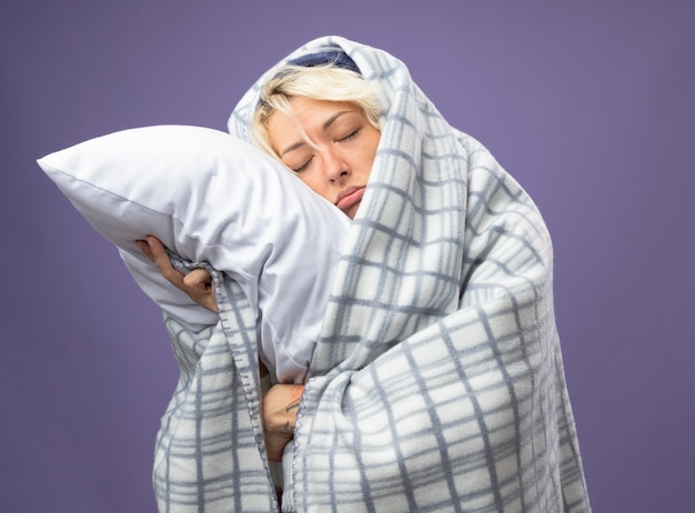 Больная нездоровая женщина с короткими волосами в теплой шапке, завернутой в одеяло, держит подушку, опираясь головой на подушку с закрытыми глазами, несчастная на фиолетовом фоне