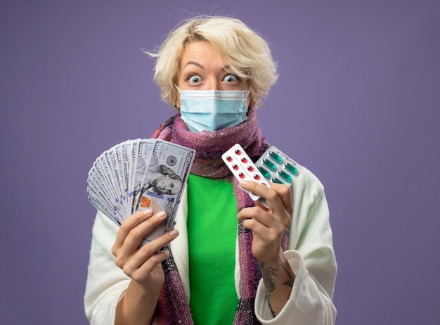 Sick unhealthy woman with short hair in warm scarf andfacial protective mask holding cash and blister with pills looking at camera being shocked standing over purple background