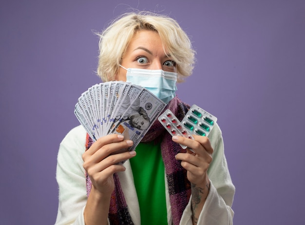 Sick unhealthy woman with short hair in warm scarf andfacial protective mask holding cash and blister with pills  worried and confused standing over purple wall