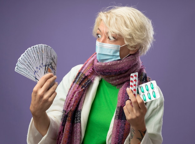 Free photo sick unhealthy woman with short hair in warm scarf andfacial protective mask holding cash and pill looking confused and worried having doubts standing over purple wall