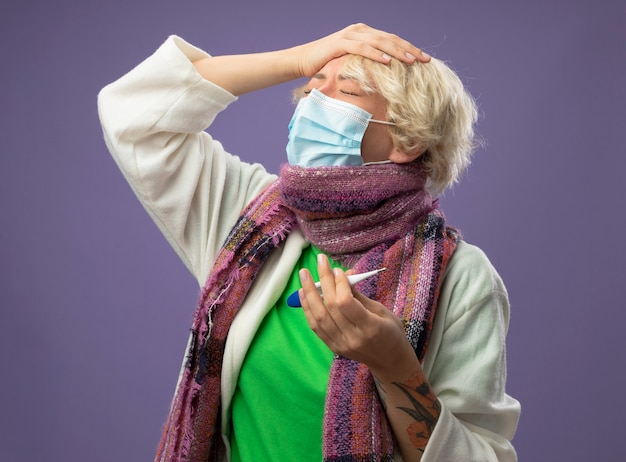 暖かいスカーフと顔の保護マスクの短い髪の病気の不健康な女性は、紫色の背景の上に立って目を閉じて彼女の頭に触れている温度計を保持しています