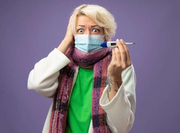 Больная нездоровая женщина с короткими волосами в теплом шарфе и защитной маске для лица, показывающая термометр, глядя в камеру в панике на фиолетовом фоне