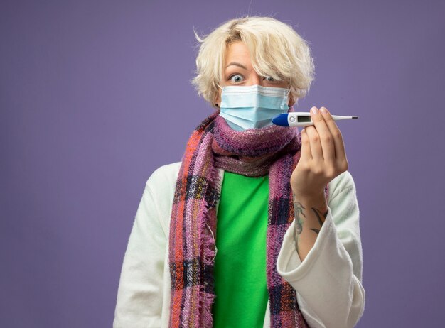 Sick unhealthy woman with short hair in warm scarf andfacial protective mask showing thermometer  in panicstanding over purple wall