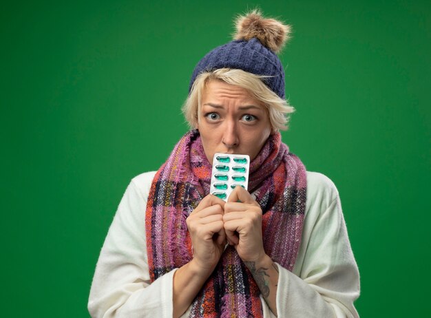 Sick unhealthy woman with short hair in warm scarf and hat feeling unwell holding blisters with pills  being upset and unhappy standing over green wall