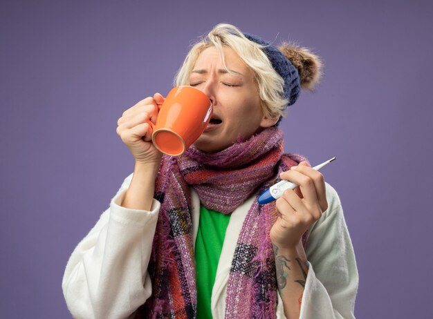 暖かいスカーフと帽子の短い髪の病気の不健康な女性は、紫色の壁の上に立ってお茶を飲みに行く熱いお茶と温度計のカップを持って気分が悪い