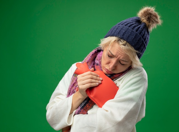 Free photo sick unhealthy woman with short hair in warm scarf and hat feeling unwell holding water bottle to kep warm standing over green background