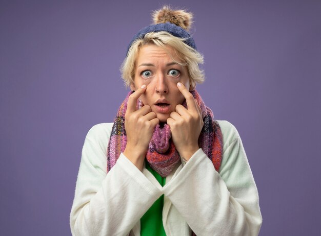 Sick unhealthy woman with short hair in warm scarf and hat feeling unwell looking at camera pointing with index figner at her eyes being surprisedover purple background