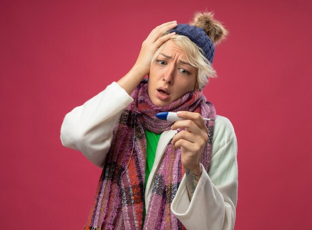 Sick unhealthy woman with short hair in warm scarf and hat feeling unwell looking at thermometer worried and confused standing over pink wall