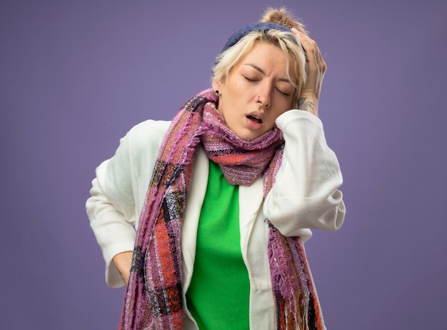 Foto gratuita donna malata malsana con i capelli corti in sciarpa calda e cappello sensazione di malessere toccando la testa che soffre di influenza e forte mal di testa su sfondo viola